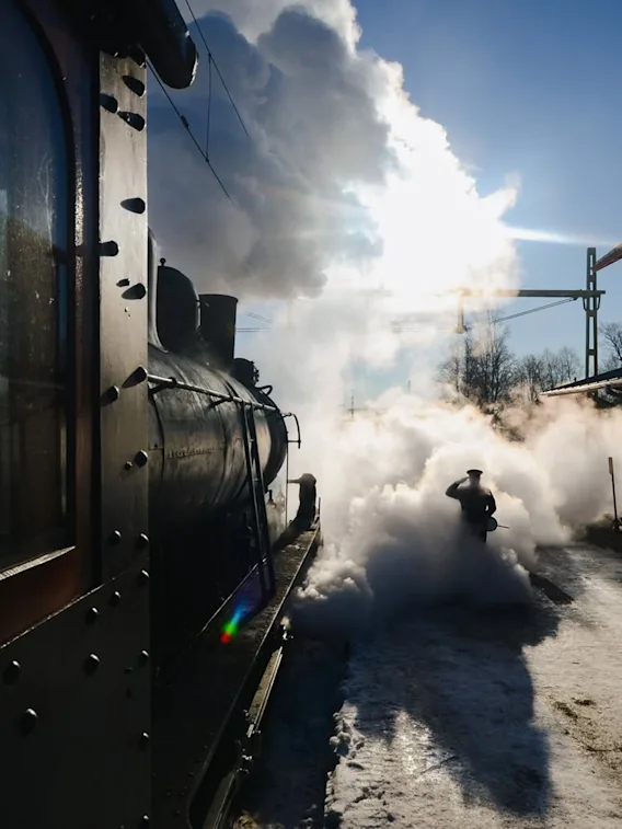 Ett ånglok på en snötäckt station, insvept i ett moln av vit ånga, med en person som går igenom det under en ljus himmel.