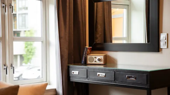 A table with drawers holds a small radio and a book, situated below a large mirror. Curtains and windows in a cozy room provide natural light.