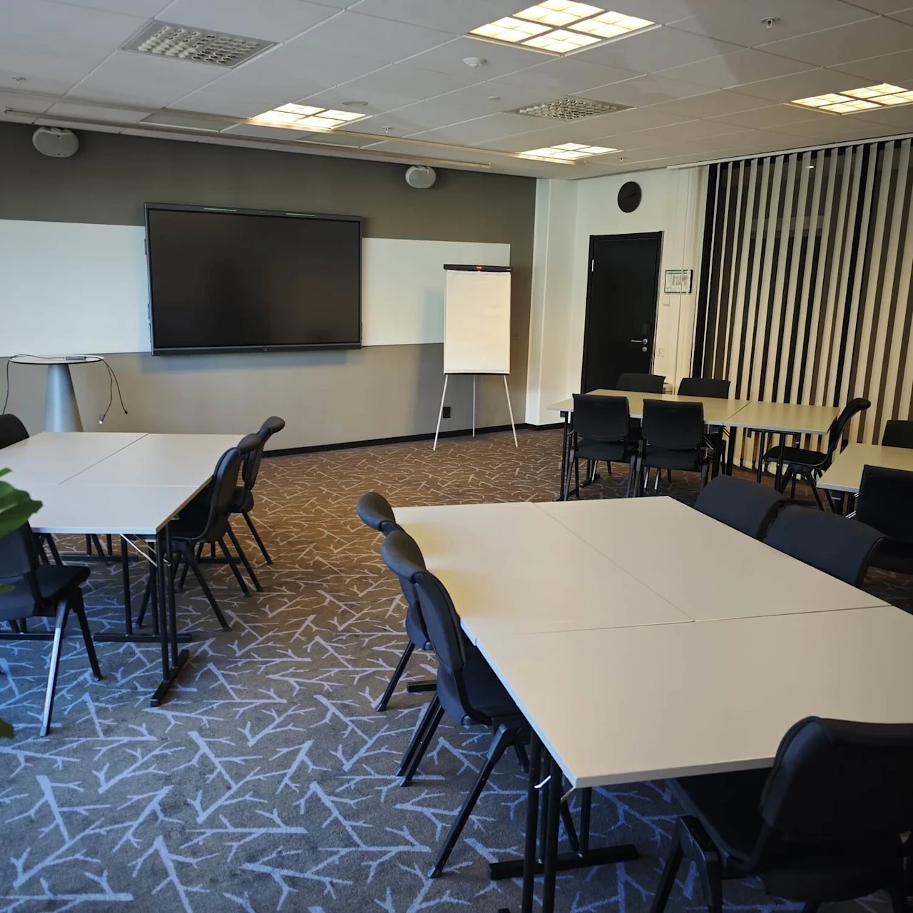 Ett konferensrum med rektangulära bord och stolar arrangerade i grupper. En stor skärm och whiteboard finns på en vägg. En krukväxt syns i förgrunden.