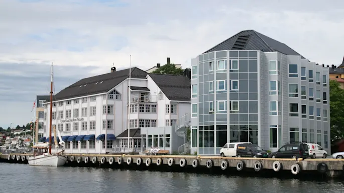 A waterside hotel with a traditional façade and a modern glass building stands by a dock. Several cars and a moored sailboat are present. Text: "Clarion Collection Hotel Tyholmen."