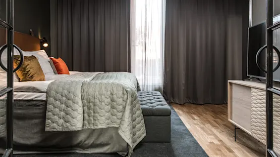 En pænt redt seng med grønt sengetøj og puder står mod en mørk væg, med et vindue dækket af grå gardiner i et hyggeligt, moderne soveværelse.