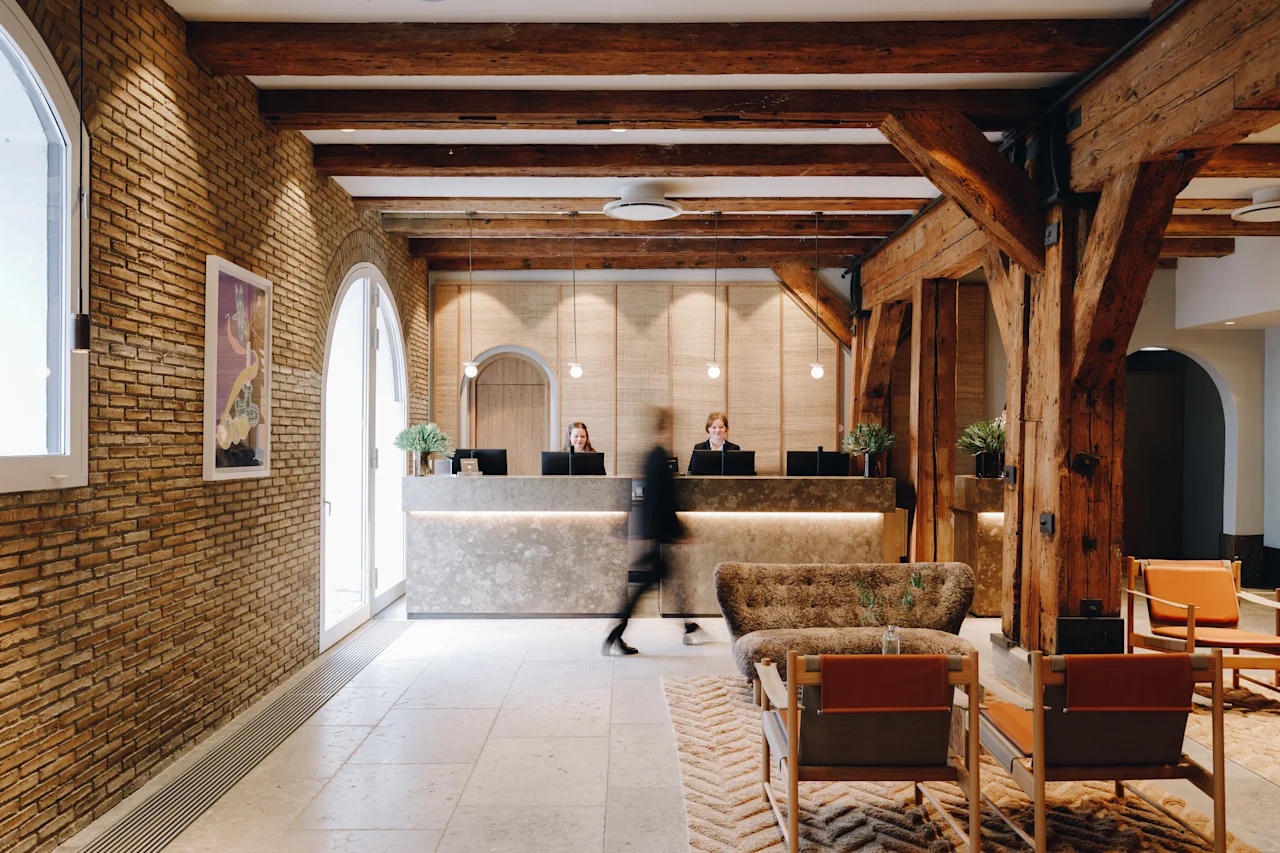 En person går gjennom en hotellobby med resepsjon, ansatte, murvegger og trebjelker, noe som skaper en varm atmosfære.