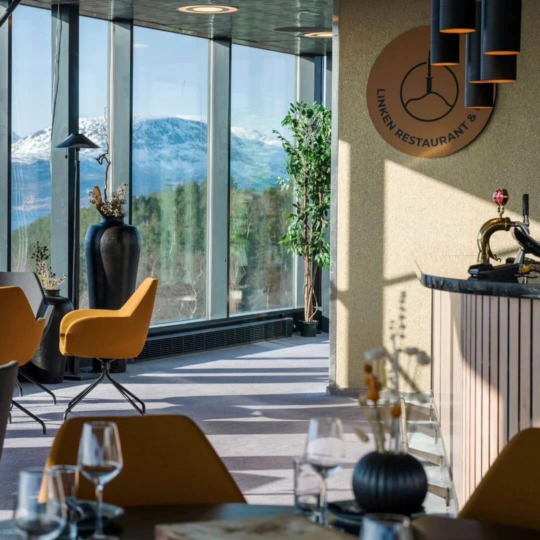 Borde og stole står i en moderne restaurant med gulv-til-loft vinduer, der tilbyder udsigt over sneklædte bjerge og vand. Et skilt lyder, "LINKEN RESTAURANT &" på væggen.