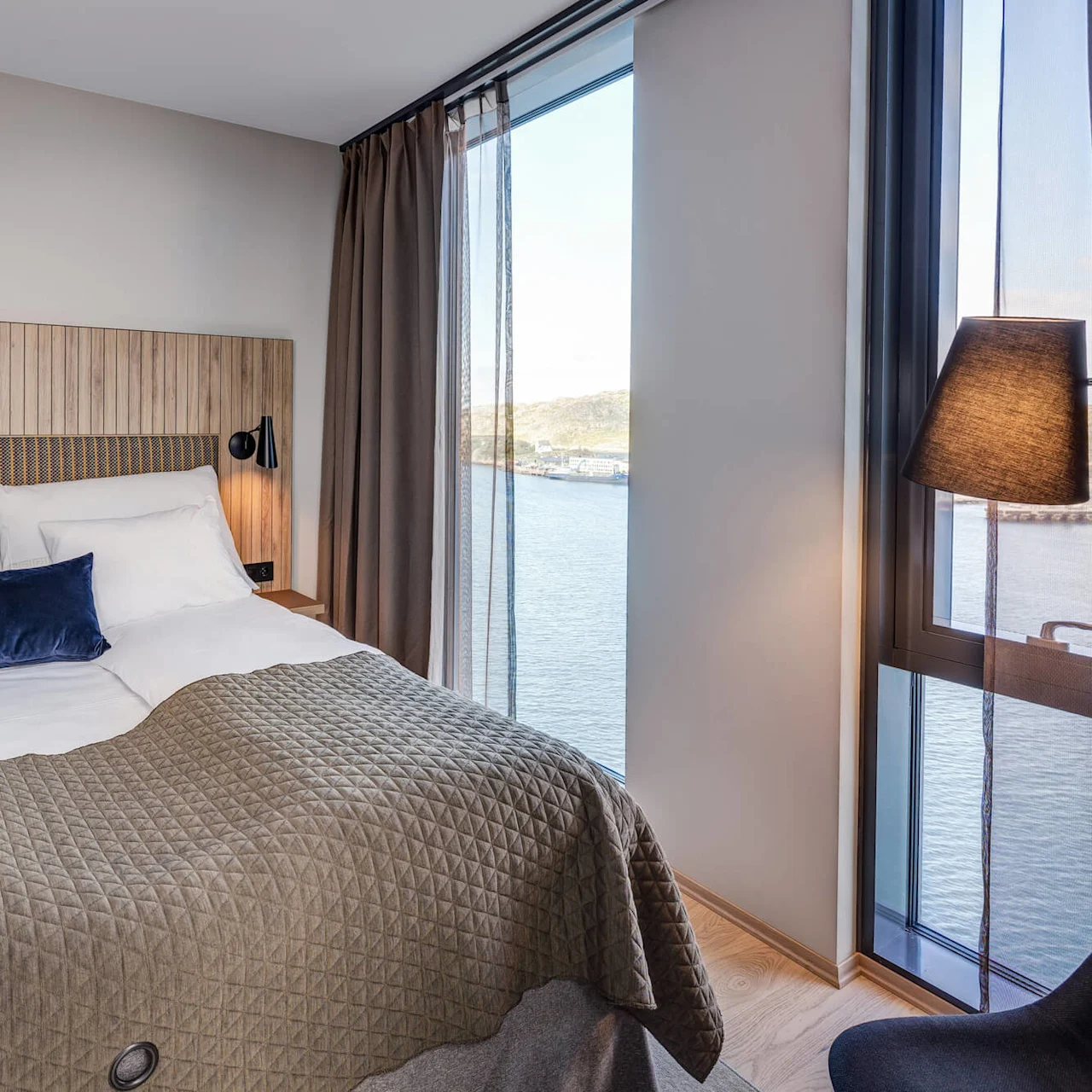 A neatly made bed with white pillows and a blue cushion, framed by wooden walls and large windows, overlooks a calm waterfront scene.