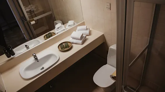A bathroom features a white sink on a beige counter with stacked towels and green glass jars. A toilet and shower are nearby. Beige tiles cover the walls.