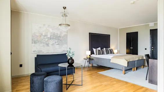 Et hyggelig hotellrom med elegant dekor. En myk seng med dekorative puter står ved siden av en sittegruppe med sofa, rundt bord og krakker. En lysekrone tilfører litt luksus.