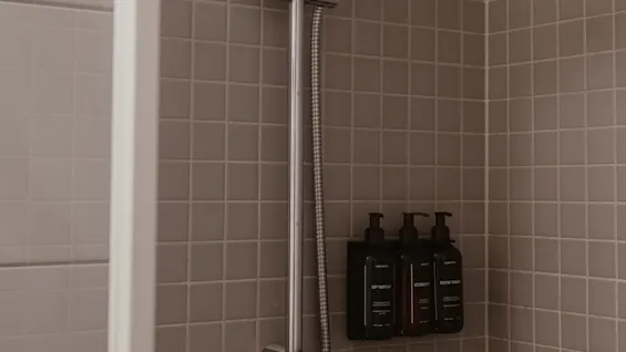 Showerhead mounted on tiled wall; three bottles labeled shampoo, conditioner, and bodywash attached beside. Context: enclosed shower cabin with neutral-colored tiles.