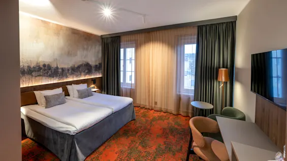 A modern hotel room with twin beds, a large artwork, curtains, a carpeted floor, a desk, chair, and a wall-mounted TV.