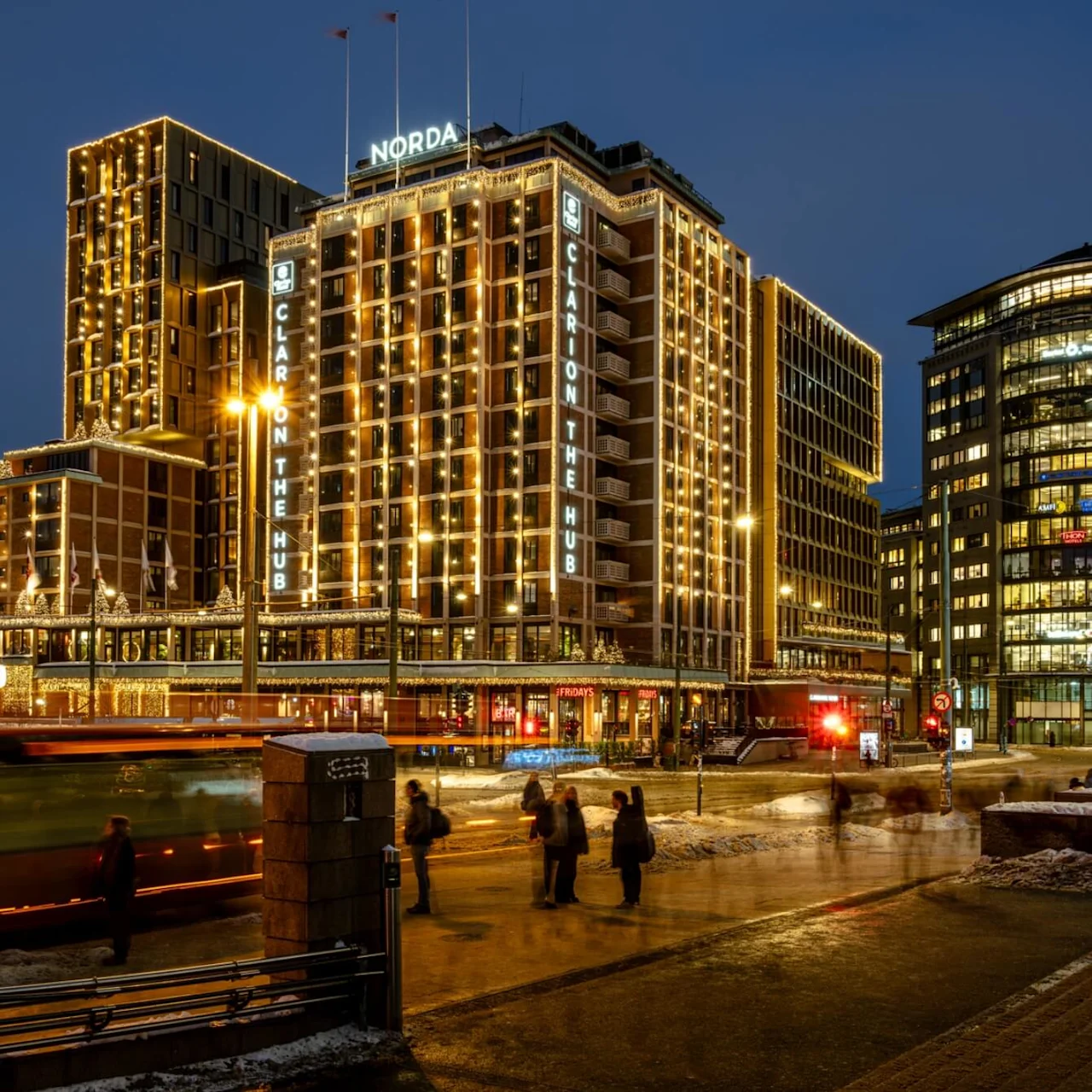 A tall, illuminated hotel building, decorated with lights, stands beside a busy street where a bus passes and people walk. Text includes: "Ruter", "Norda", "Clarion The Hub", "TGI Friday's".