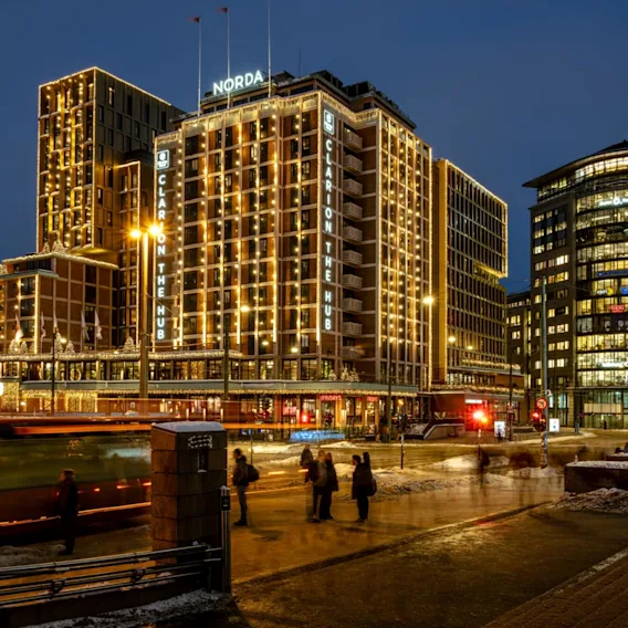 A tall, illuminated hotel building, decorated with lights, stands beside a busy street where a bus passes and people walk. Text includes: "Ruter", "Norda", "Clarion The Hub", "TGI Friday's".