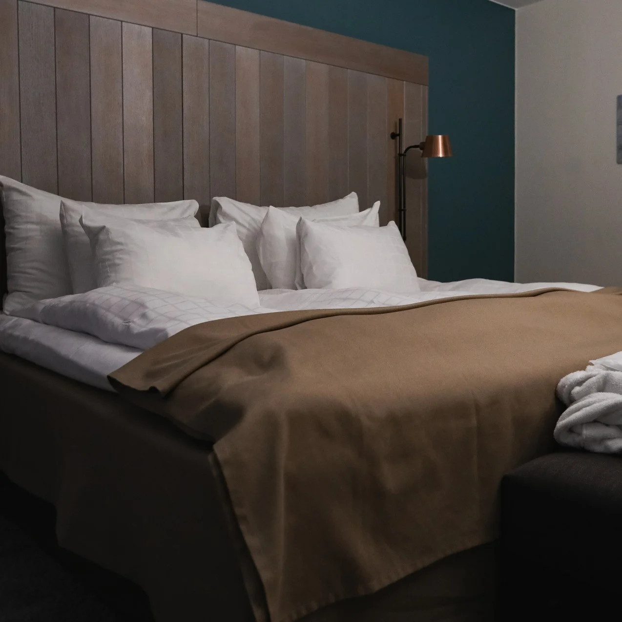 A neatly made bed with white pillows and a brown blanket sits beneath a wooden headboard. Two robes lie on a bench, in a softly lit room with teal and cream walls.