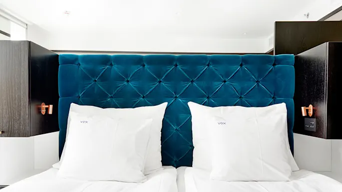 To pent oppredde hvite senger med "VOX" broderte puter hvilende mot en dypt blå, knappetrukket sengegavl i et mykt belyst rom, flankert av mørke trepaneler.