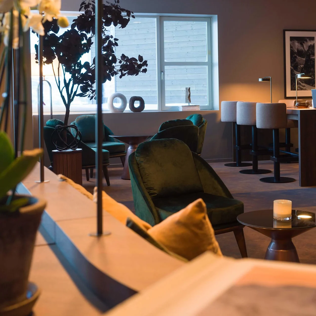 A person works on a laptop at a wooden table in a cozy, warmly-lit lounge area, surrounded by plush green chairs, plants, and modern decor.
