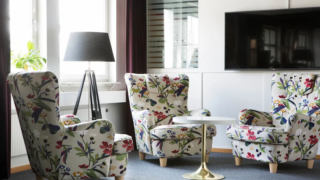 To lænestole med blomstermønstre befinder sig i en stue, omkranser et lille rundt bord, tæt på en lampe og et vægmonteret fjernsyn.