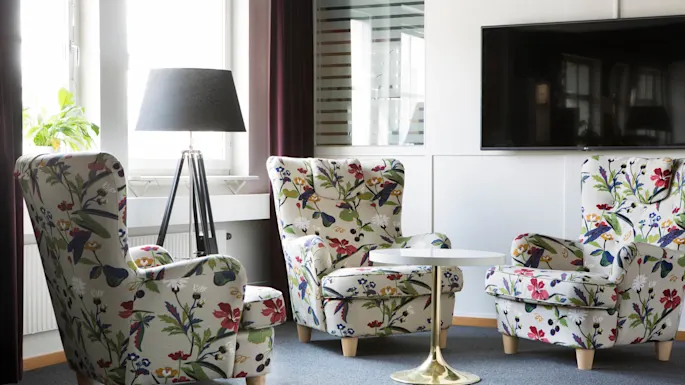 To lenestoler med blomstermønster står i en stue, flankerer et lite rundt bord, med en lampe og en veggmontert TV i nærheten.