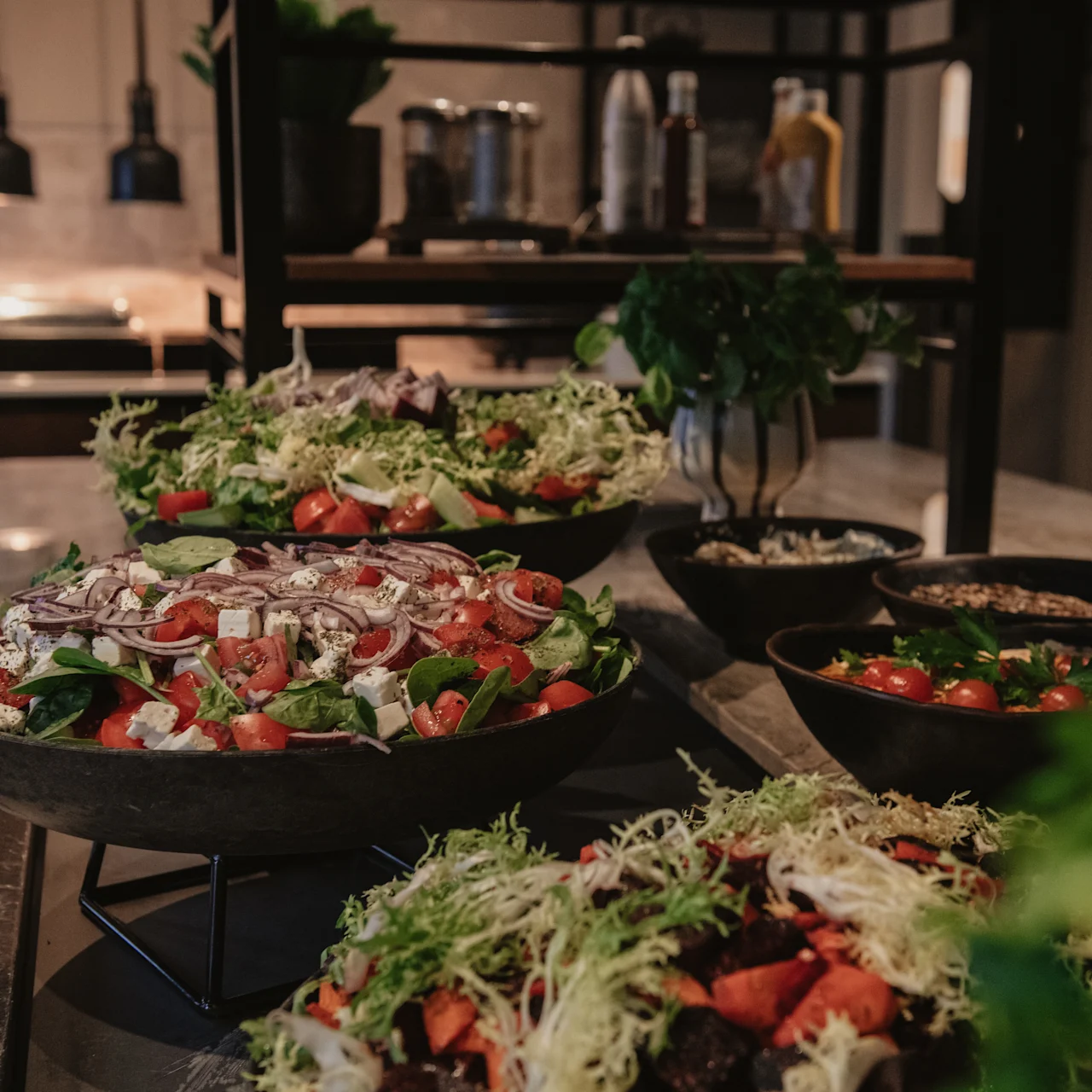 Large bowls of colorful salads are displayed on a kitchen counter, featuring fresh greens, tomatoes, and various vegetables, under warm ambient lighting.