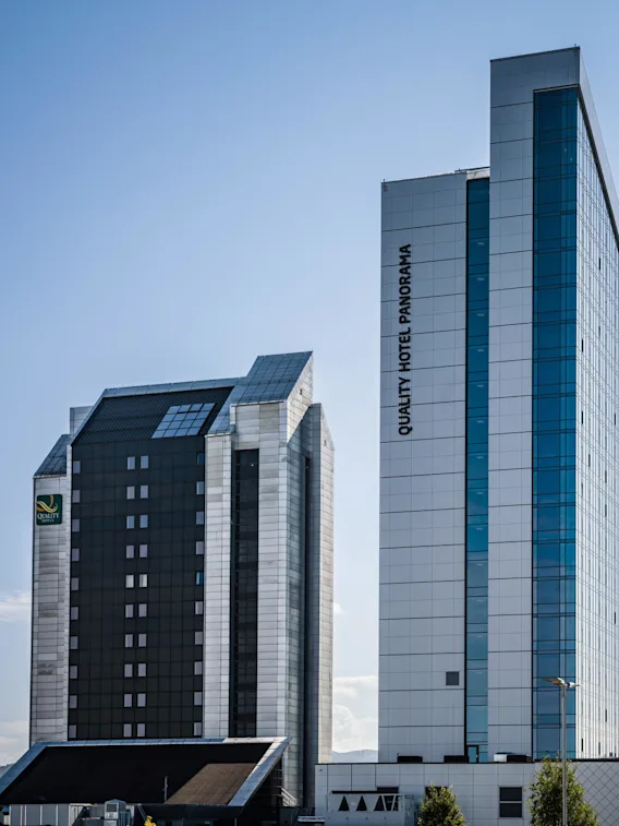 To høye bygninger står nær hverandre under en klarblå himmel. En har teksten "QUALITY HOTEL PANORAMA" vertikalt. Den urbane settingen har lave åser i bakgrunnen.