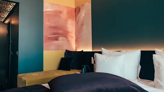 A bed with white pillows is placed against a dark wall, adjacent to a yellow bench. Abstract pink artwork decorates the wall. 
