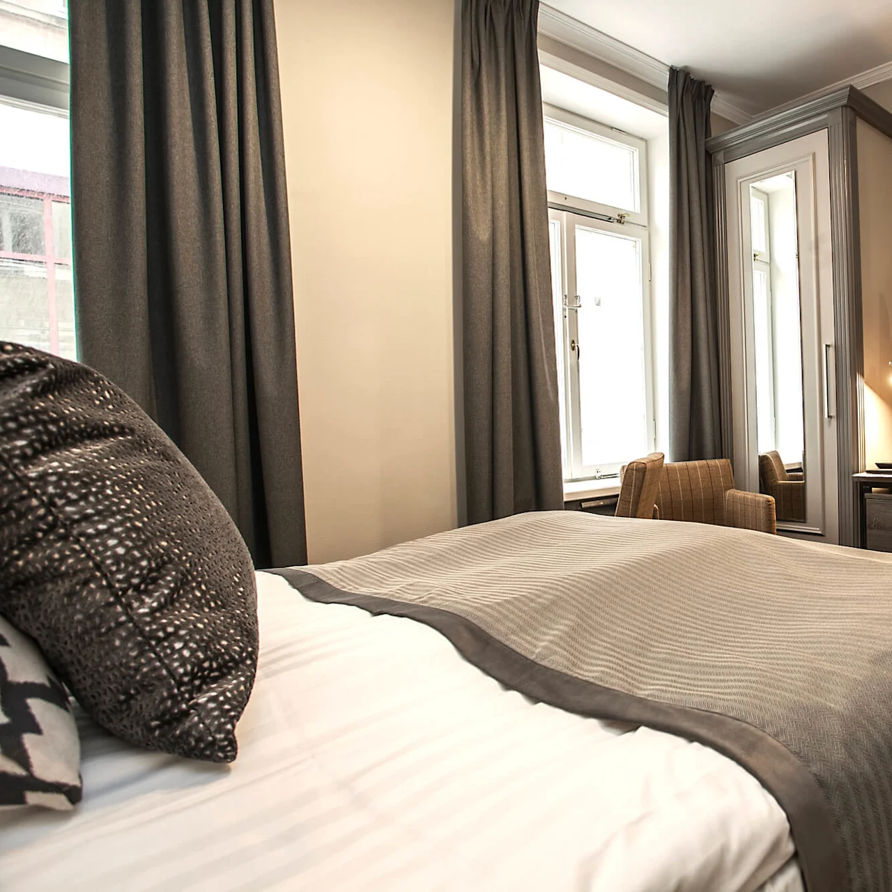 En ryddig seng med dekorative puter i et lyst hotellrom med skrivebord, stol og TV.
