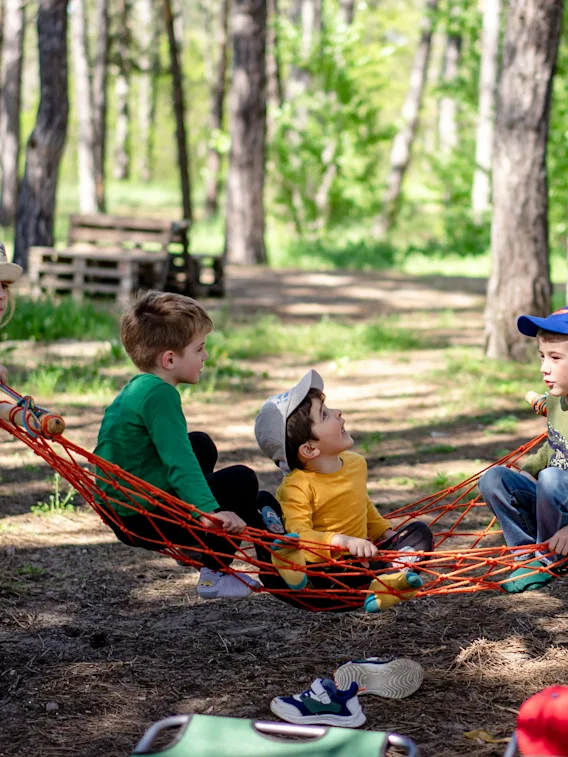 Barn leker i en hengekøye i en solrik skog. Ett barn dytter hengekøyen mens andre sitter og prater.
