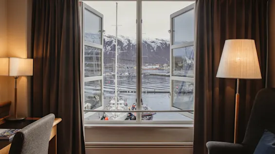 A cozy room with an open window offering a stunning view of a harbor with boats and snow-capped mountains.