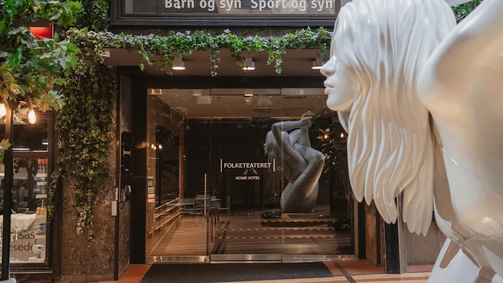Building entrance with green plants. Inside, a statue in a yoga pose. Text above door: Barn og syn Sport og syn.