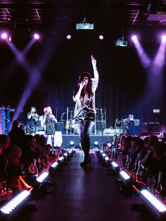 A singer performs energetically on a stage runway, surrounded by an audience. Backing singers and a band play under purple stage lights in a lively concert atmosphere.