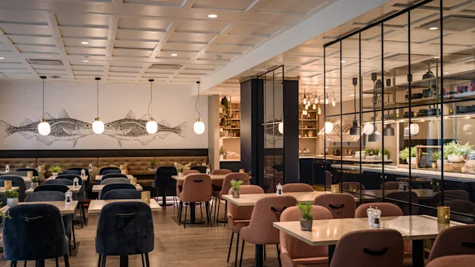 A bright and inviting restaurant with many empty tables and chairs, decorated with fish art and a sleek glass-paneled area.