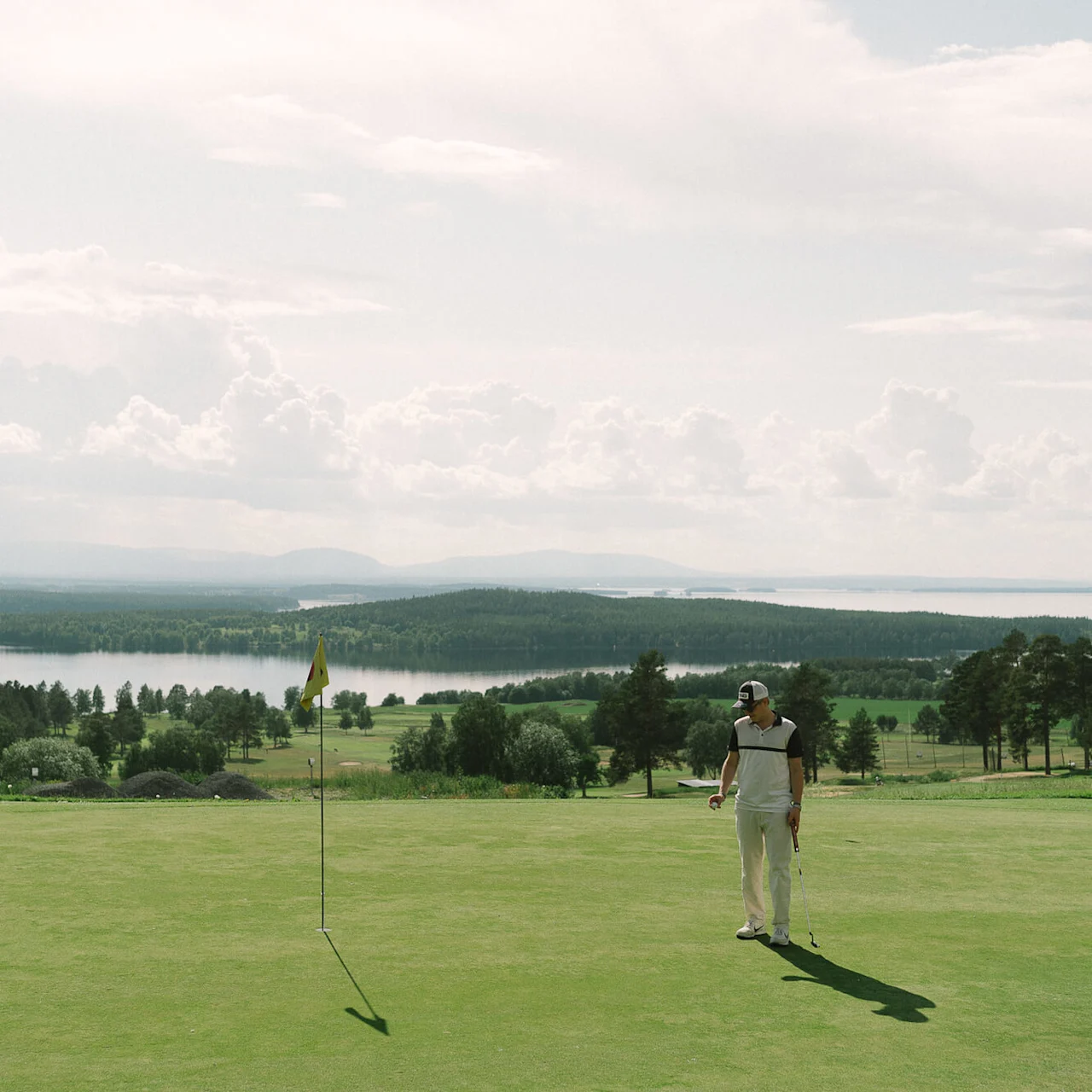 Golfspelare står på en grön bana, håller en klubba, tittar mot en flagga. I bakgrunden syns ett rofyllt landskap med avlägsna träd och en sjö under en molnig himmel.
