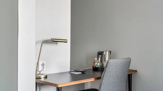 A desk with a lamp, phone, kettle, and chair, against a two-tone wall.