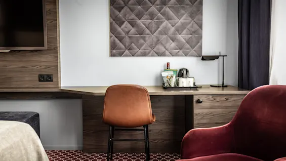 Ett mysigt hotellrum med en elegant skrivbords- och stolsuppsättning, en modern TV på väggen och en bekväm, mörkröd fåtölj. En bricka med en vattenkokare och koppar står på skrivbordet, vilket ger en inbjudande känsla.