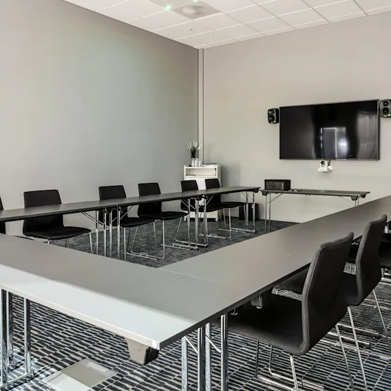 U-shaped conference table with black chairs, facing a wall-mounted screen and two speakers, in a modern meeting room with grey walls and patterned carpet.