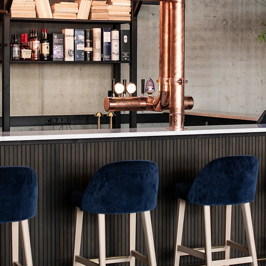 En moderne bar med bløde blå barstole, en fremtrædende kobberølhane og hylder fyldt med drikkevarer og bøger.