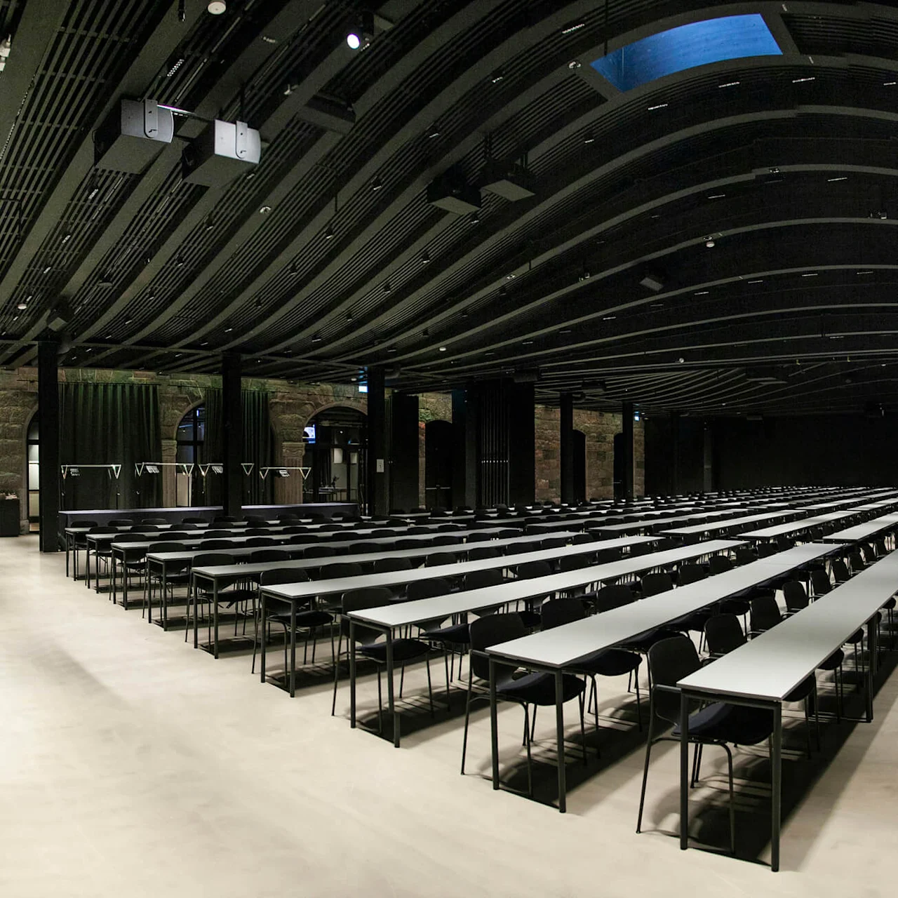 Long tables with chairs are arranged in rows facing a podium in a spacious, dark-ceilinged conference room. The walls are lined with windows and curtains under ambient lighting.