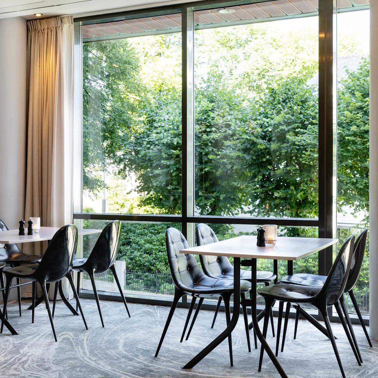 Tables with black chairs are arranged in a modern dining area, featuring large windows overlooking green trees outside. Minimalist decor with pepper shakers and small candleholders adorns the tables.