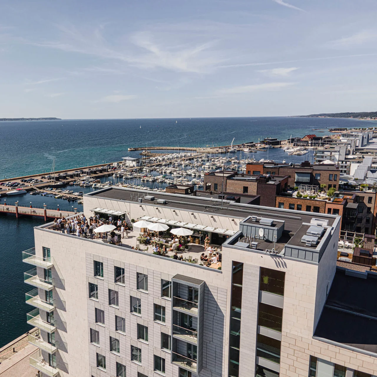 En takterrasse med mennesker under hvite parasoller, som ser over en marina fylt med båter. Scenen er satt av et stort hav og bybygninger under en klar, blå himmel.