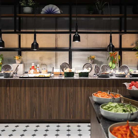 Buffet-oppstilling med ulike retter på en benkeplate, inkludert salater og sauser. Hengelamper over lyser opp trebenken i et moderne spisested med planter og dekor.