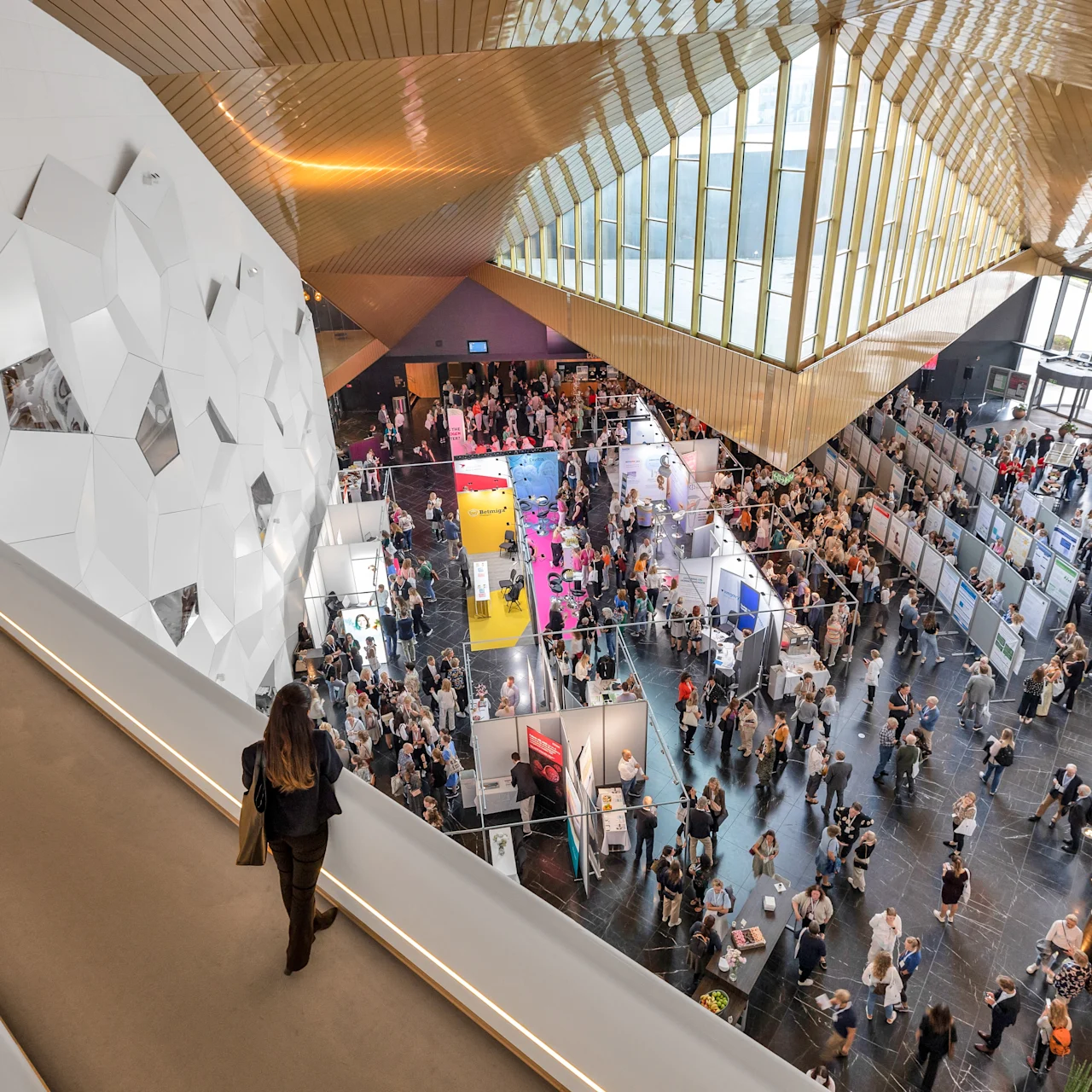 A woman walks along a balcony overlooking a busy convention hall filled with people attending. The hall features geometric wall designs and a wooden ceiling in a modern architectural setting.