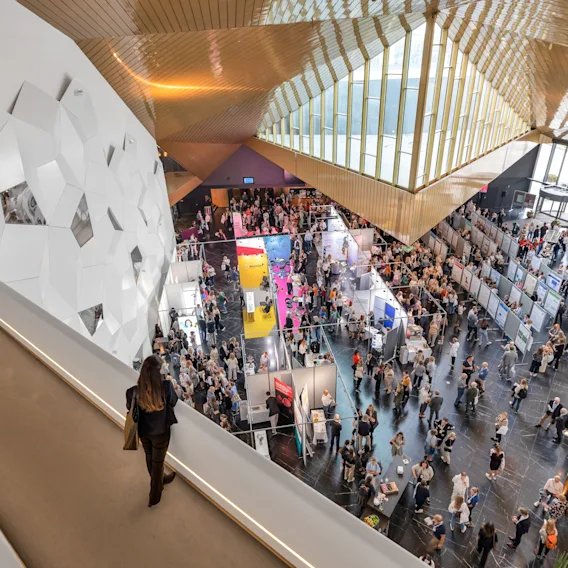 En kvinne går langs en balkong med utsikt over en travel messehall fylt med mennesker. Hallen har geometriske veggdesign og et tretak i et moderne arkitektonisk miljø.