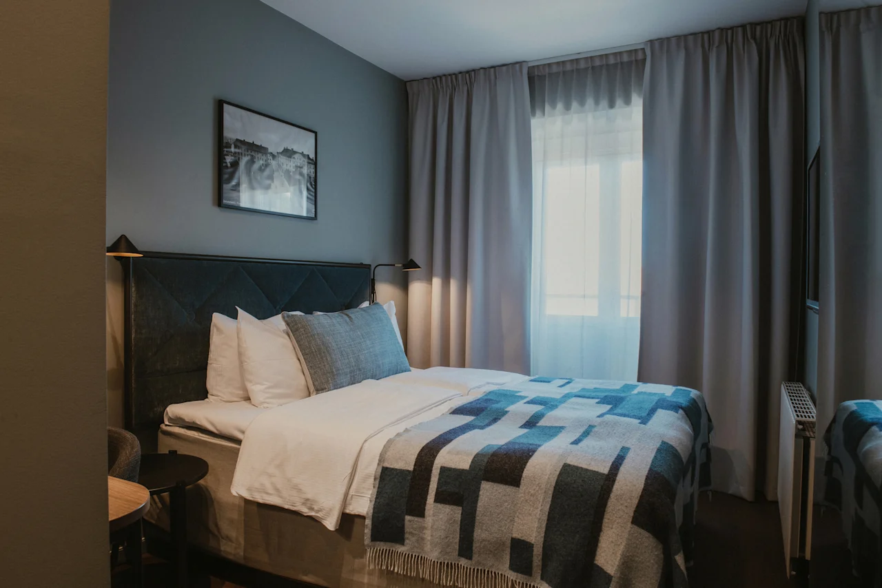 Ett inbjudande hotellrum med en säng, mönstrad filt och mjuka kuddar. Ljus strömmar in genom fönstret och skapar en lugn atmosfär.