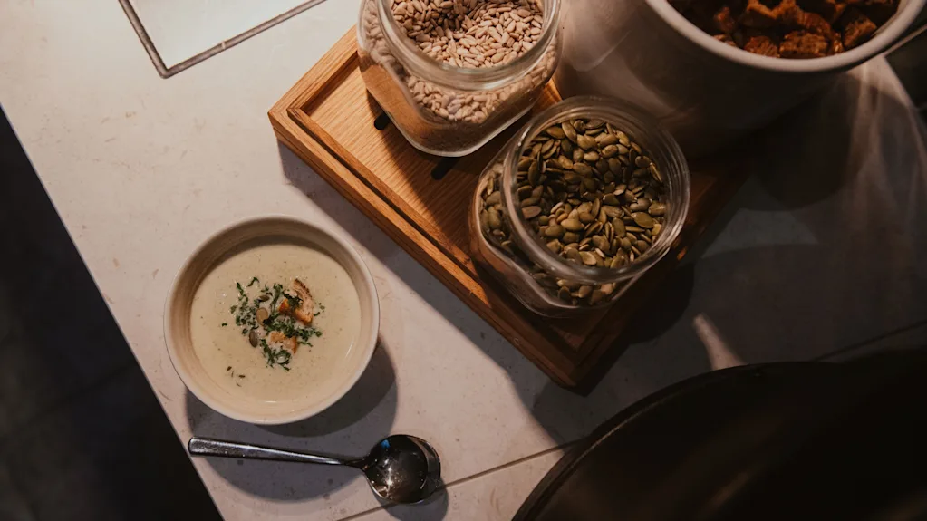 En krämig soppa i en skål med en sked, bredvid burkar med frön och krutonger, redo för en utsökt måltid.