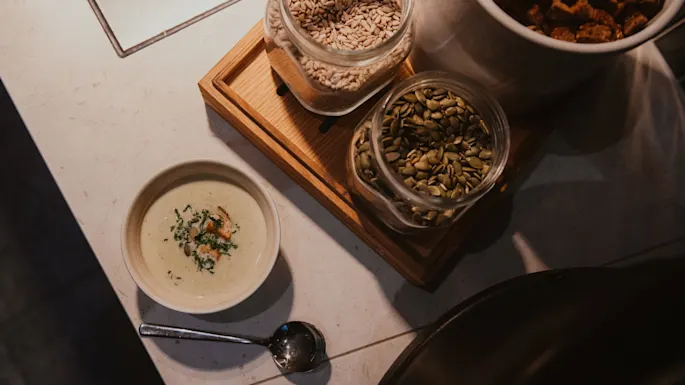 En kremet suppe i en bolle med en skje, ved siden av glass med frø og krutonger, klar for et deilig måltid.