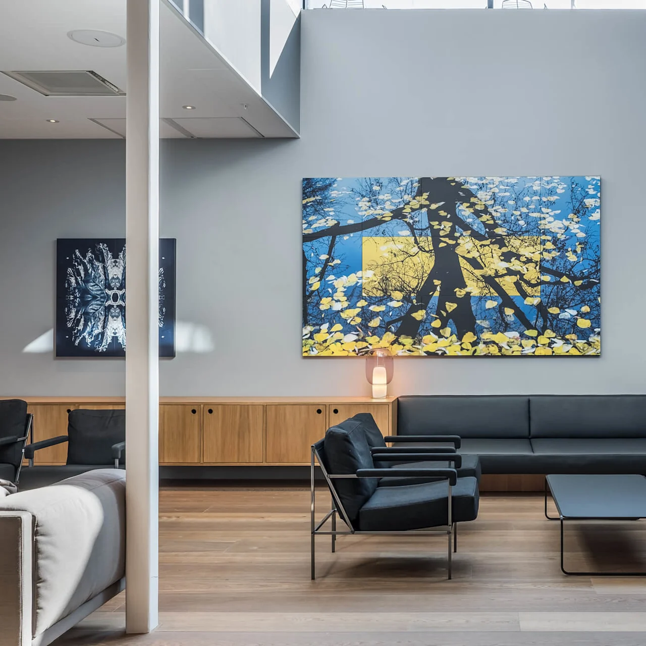 Modern living room featuring black sofas and armchairs facing a minimalist coffee table. Large abstract artwork with blue and yellow tones adorns the wall, surrounded by a spacious, well-lit interior.