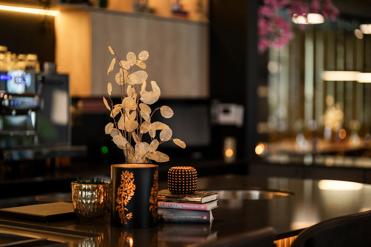 En koselig borddekking i en restaurant eller bar, med en dekorativ plante i en svart og gullfarget vase, en liten gullpotte og bøker.