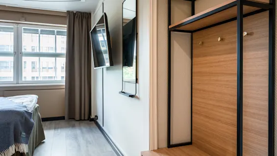 A wall-mounted flat-screen TV facing a neatly made bed with blue blankets in a modern, minimalist hotel room featuring large windows and a wooden coat rack with shelves.