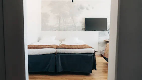 A bright hotel room featuring two single beds made side by side, wooden flooring, and a wall-mounted TV.