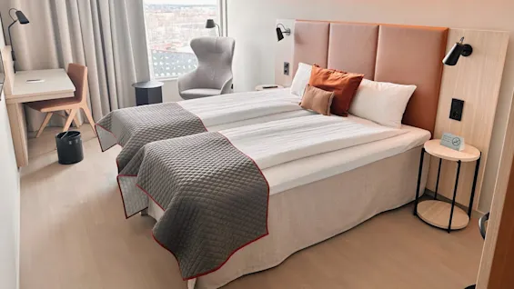 A tidy hotel room features a neatly made double bed with gray and orange pillows, accompanied by a work desk, chair, and armchair by a window. No smoking sign present.