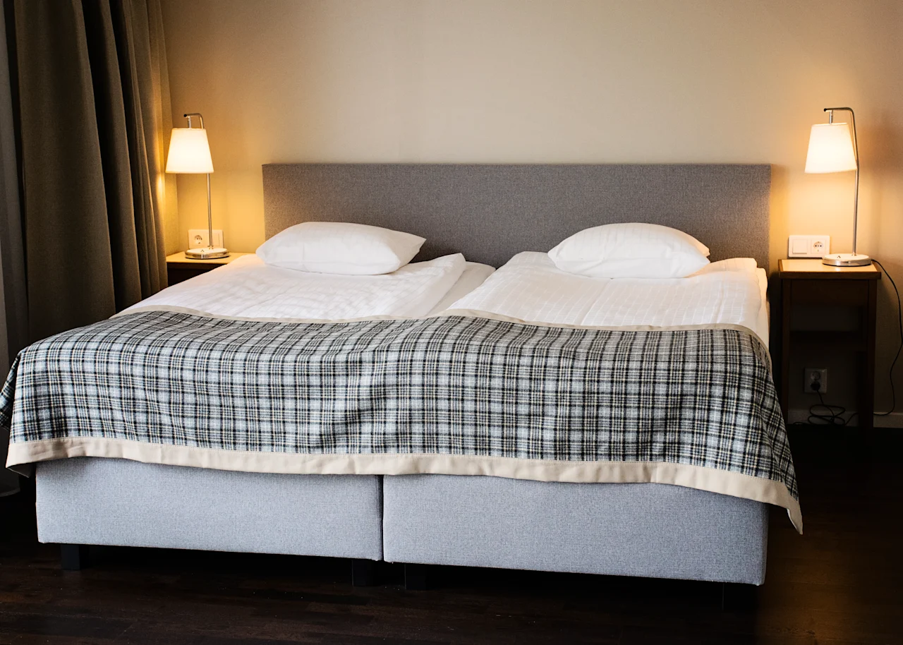 Viihtyisä hotellihuone, jossa kaksi sänkyä, ruudullinen peitto ja yöpöytälamput, valmiina mukavaan oleskeluun.