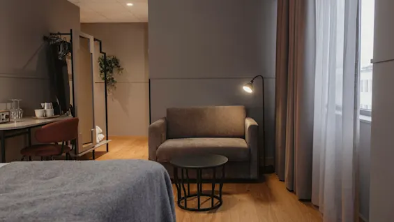 Ett mysigt hotellrum med en säng, en bekväm fåtölj, ett skrivbord och ett fönster med naturligt ljus.