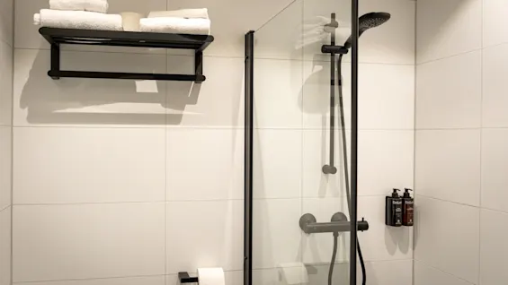 A clean, modern bathroom with white tiled walls, a shower with black fixtures, and fresh white towels on a rack.